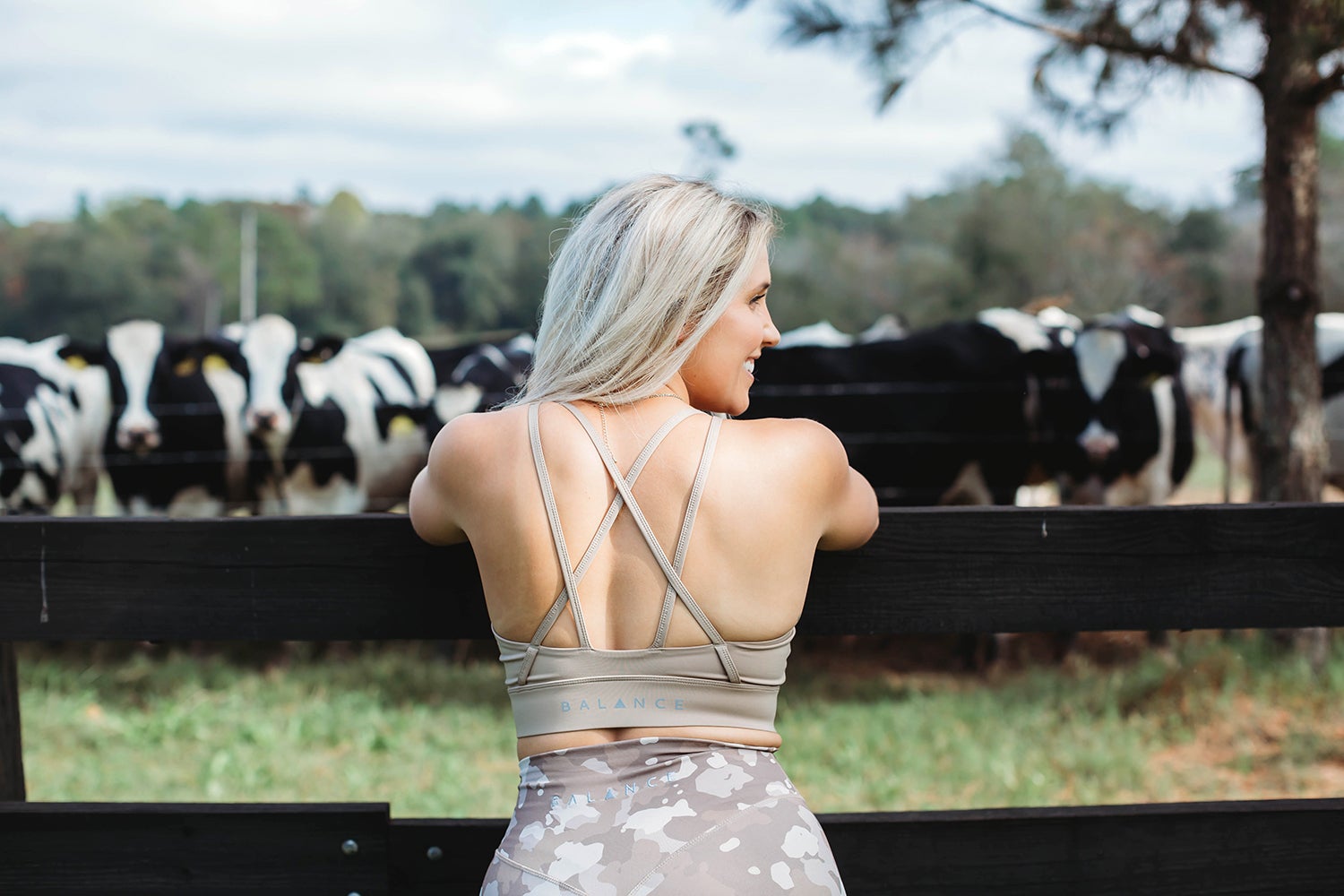 A woman in beige exercise clothing facing towards a herd of Holstein cows in a field
