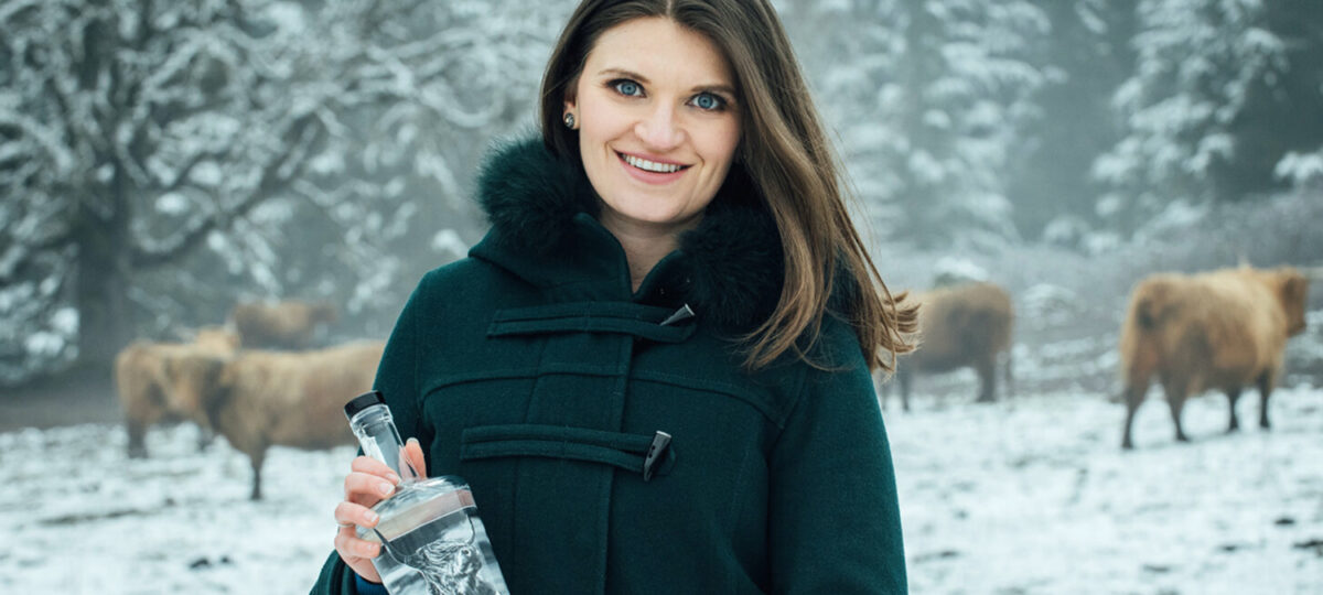 A woman with a black coat holds a bottle of clear liquor against a snowy backdrop