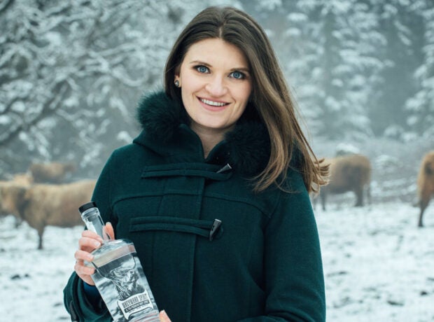A woman with a black coat holds a bottle of clear liquor against a snowy backdrop