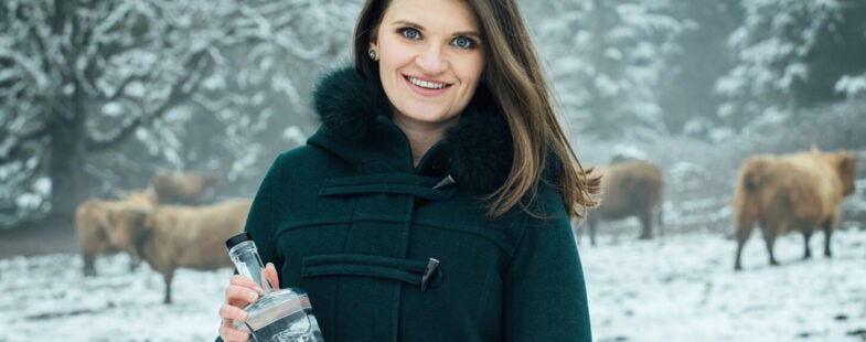 A woman with a black coat holds a bottle of clear liquor against a snowy backdrop