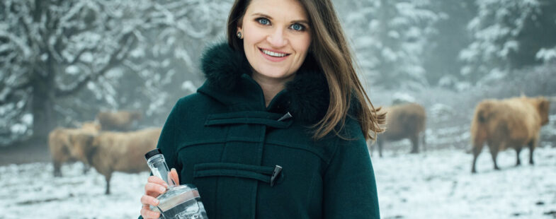 A woman with a black coat holds a bottle of clear liquor against a snowy backdrop