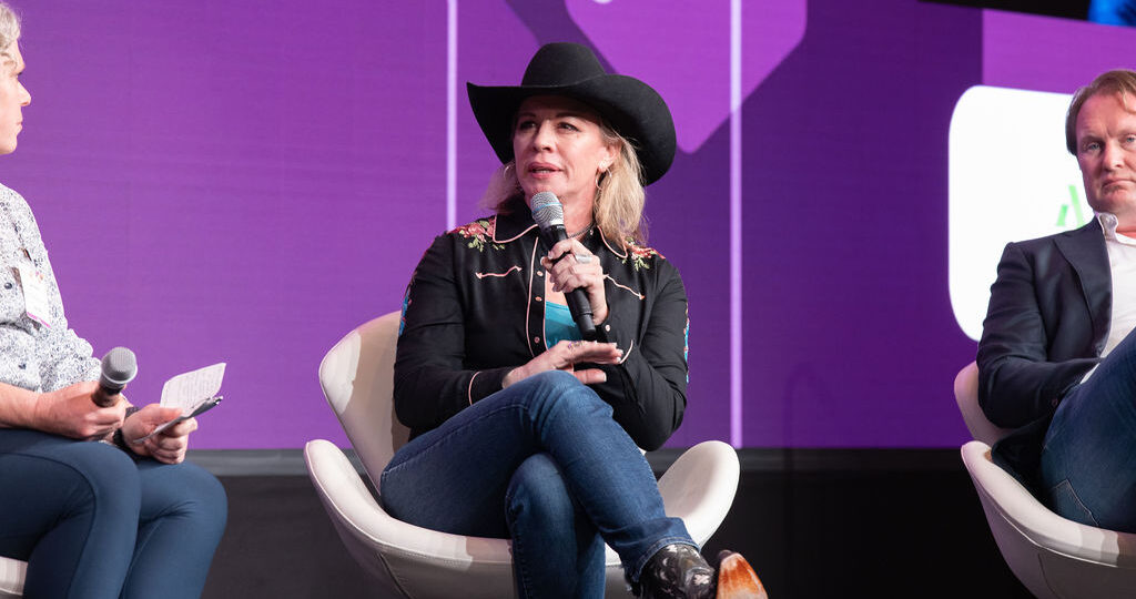 A woman in a cowboy hat speaks into a microphone on a stage, flanked by two men