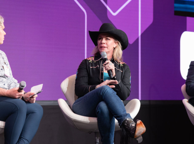 A woman in a cowboy hat speaks into a microphone on a stage, flanked by two men