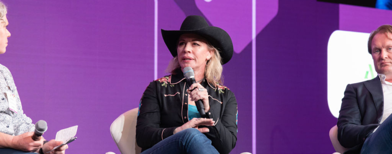 A woman in a cowboy hat speaks into a microphone on a stage, flanked by two men