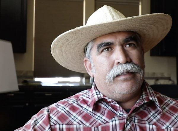 A man wearing a red shirt and straw hat talks to someone off camera