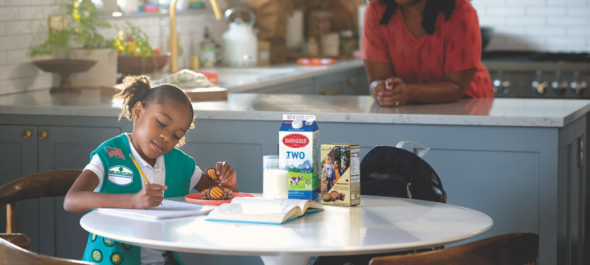 A Girl Scout sits at a kitchen table with her mom in the background