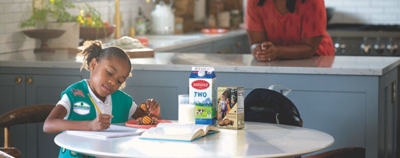 A Girl Scout sits at a kitchen table with her mom in the background