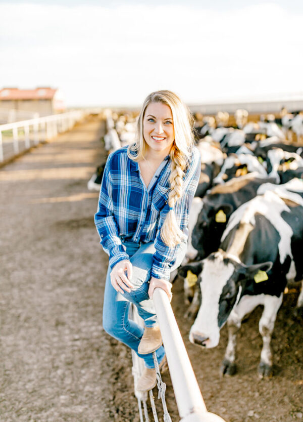 A blonde woman with a blue plaid shirt poses in front of cows