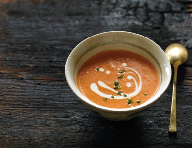 A small bowl of tomato soup displayed on a slab of wood with a brass spoon